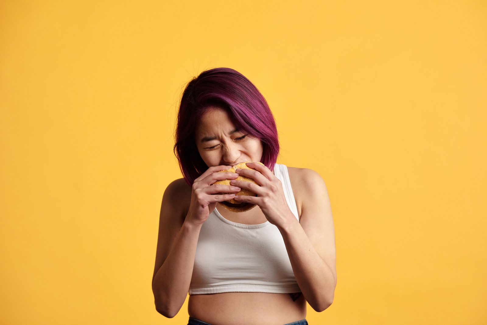 A woman with magenta hair, eyes shut, biting into a burger against a yellow background